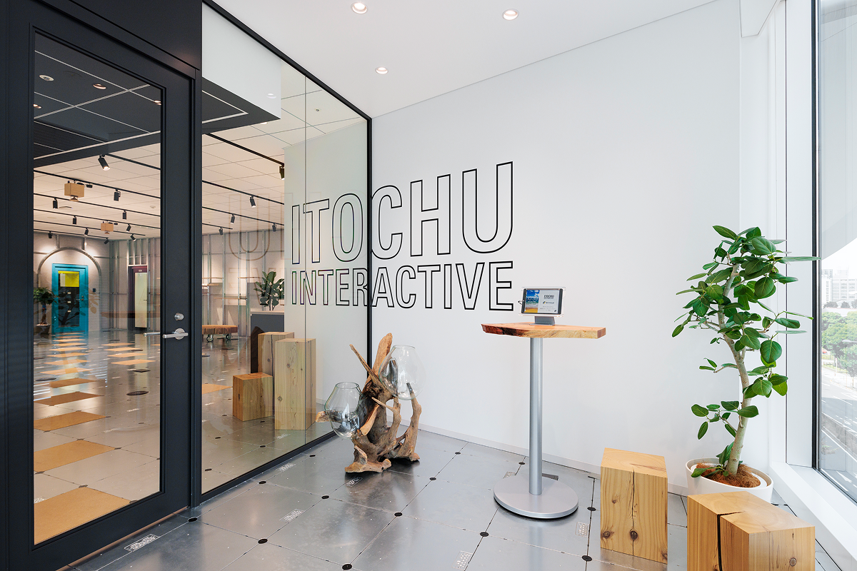写真：IICのロゴが壁に大きく描かれた明るい受付エリア。観葉植物や木製スツール、ガラスのオブジェなどが置かれている。