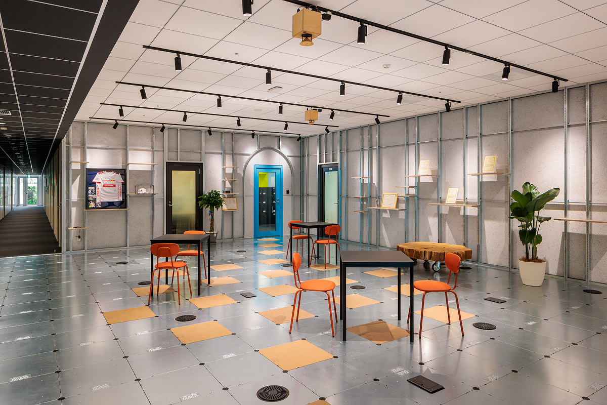 写真：四角いタイル状の床に移動可能なテーブルと椅子、観葉植物や賞状が飾られている。