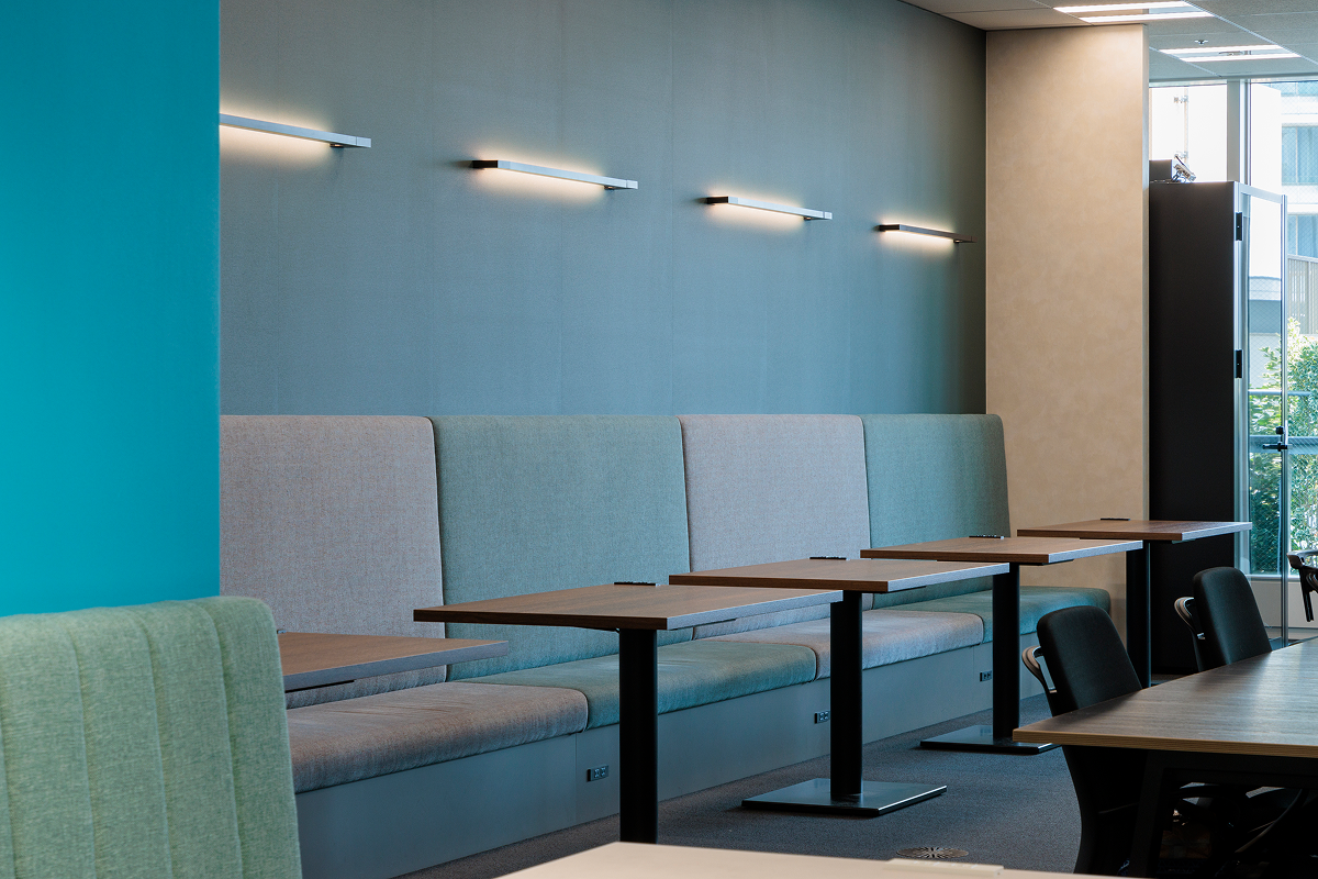 写真：壁沿いにカフェ風の長いベンチ席とテーブルが並ぶ、落ち着いた色調の空間。