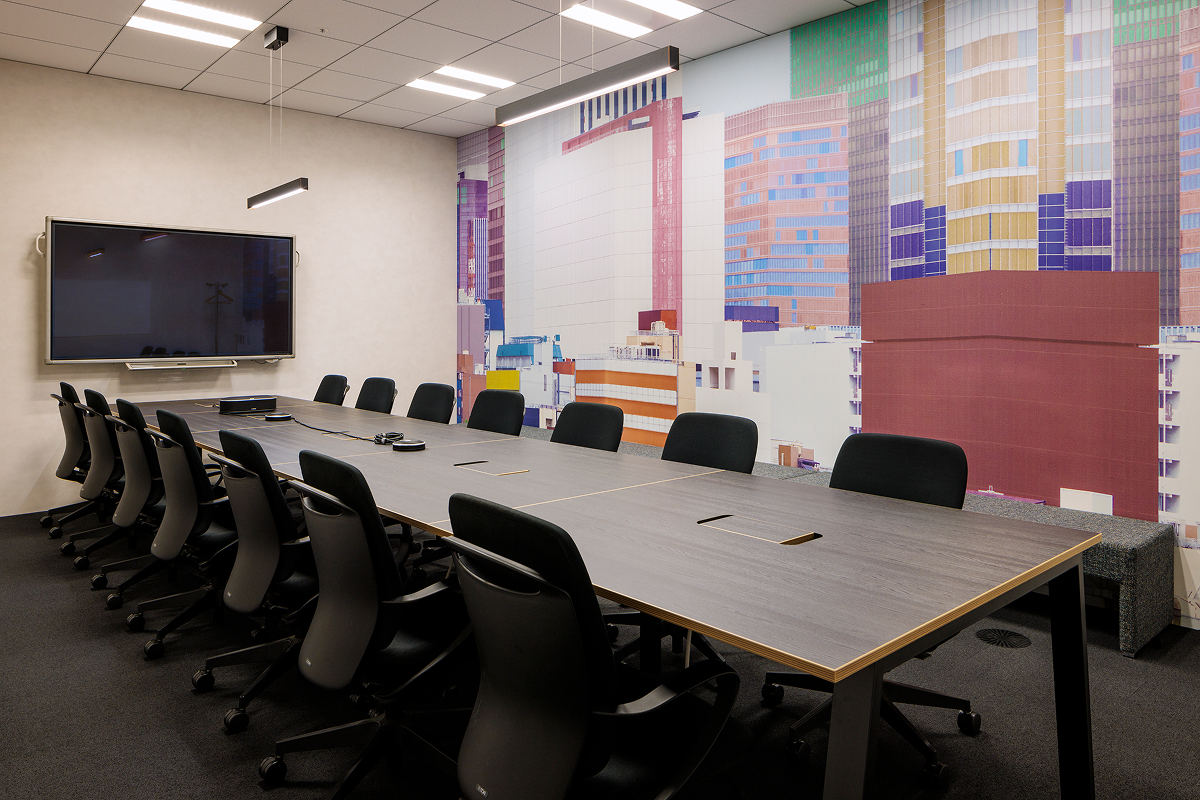 写真：壁にカラフルなビルのアートが描かれた会議室。長いテーブルと多数の椅子、壁には大型モニターが設置されている。